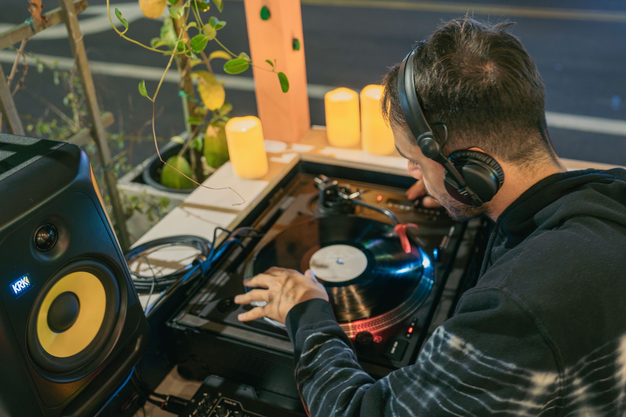 A man wearing headphones plays vinyl records on a turntable, adjusting the controls. Candles and a speaker are in the background.