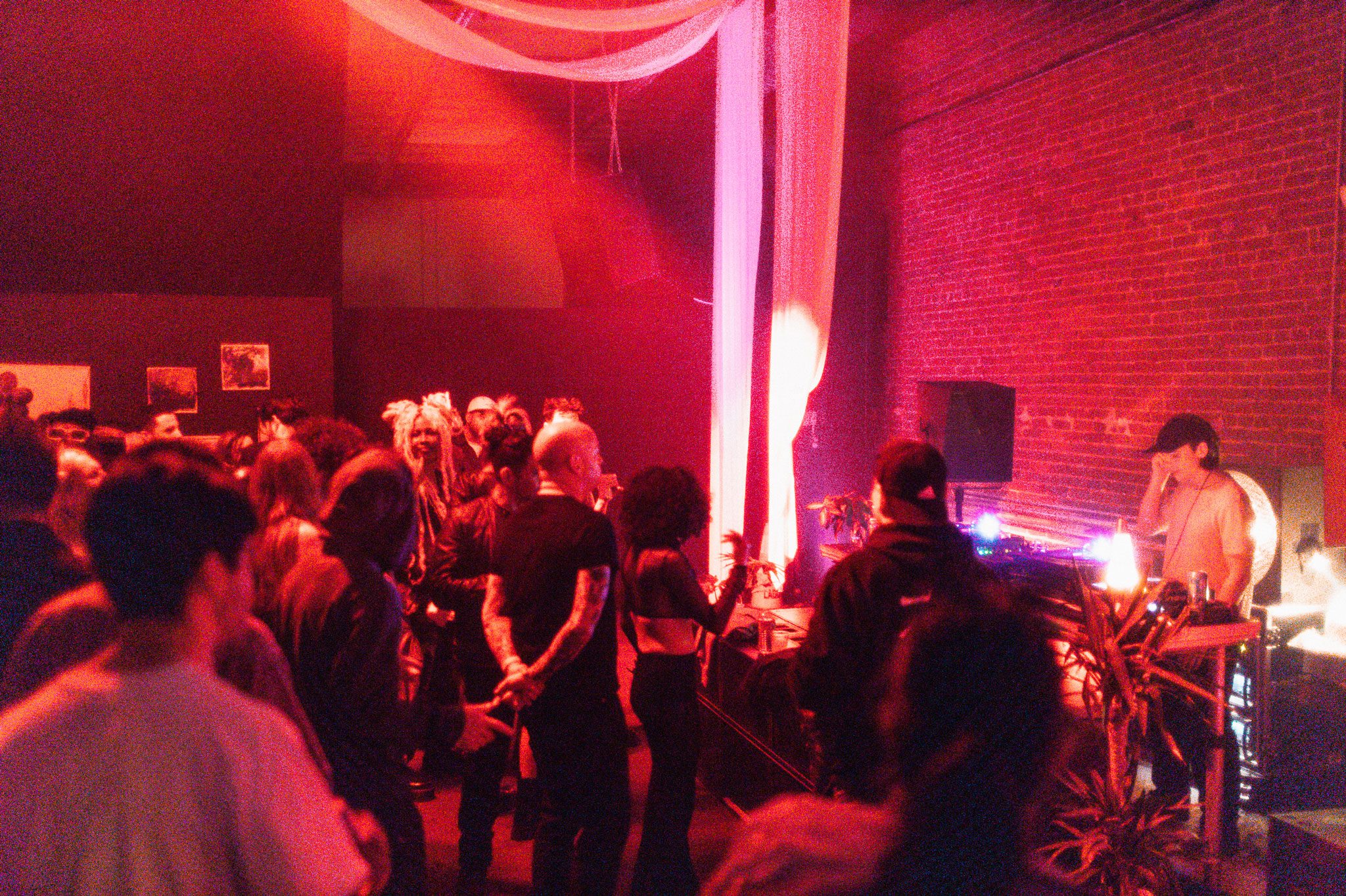 A crowd gathers in a dimly lit room with red lighting, facing towards a DJ playing music on a console, while suspended decorations hang from the ceiling.