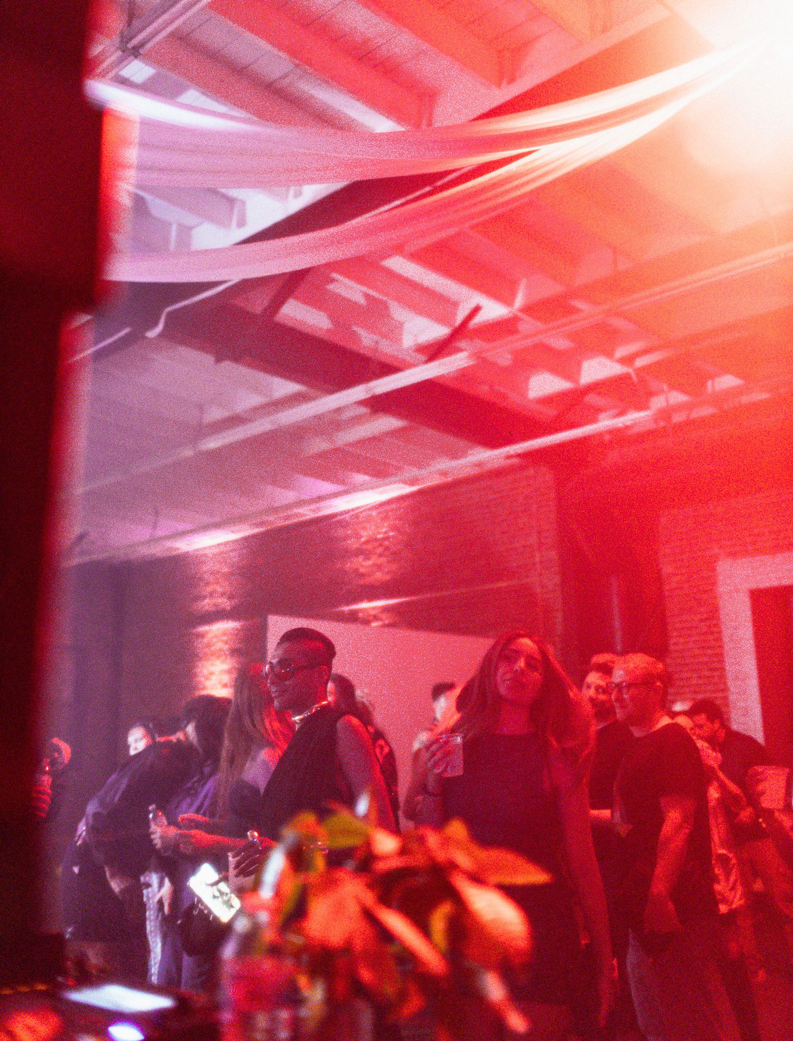 People gathered in a dimly lit room with red lighting, some holding drinks, and engaging in conversation.